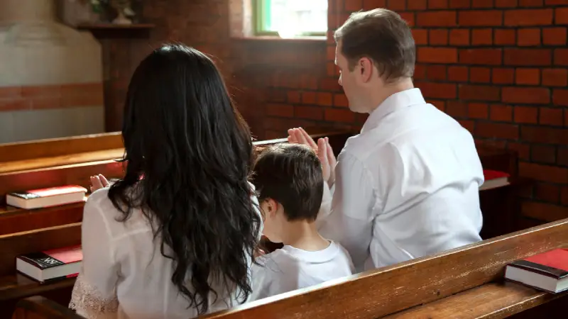 A family of three sits in wooden pews, hands clasped in prayer, surrounded by a brick church interior and floral decorations A family of three sits in wooden pews, hands clasped in prayer, surrounded by a brick church interior and floral decorations